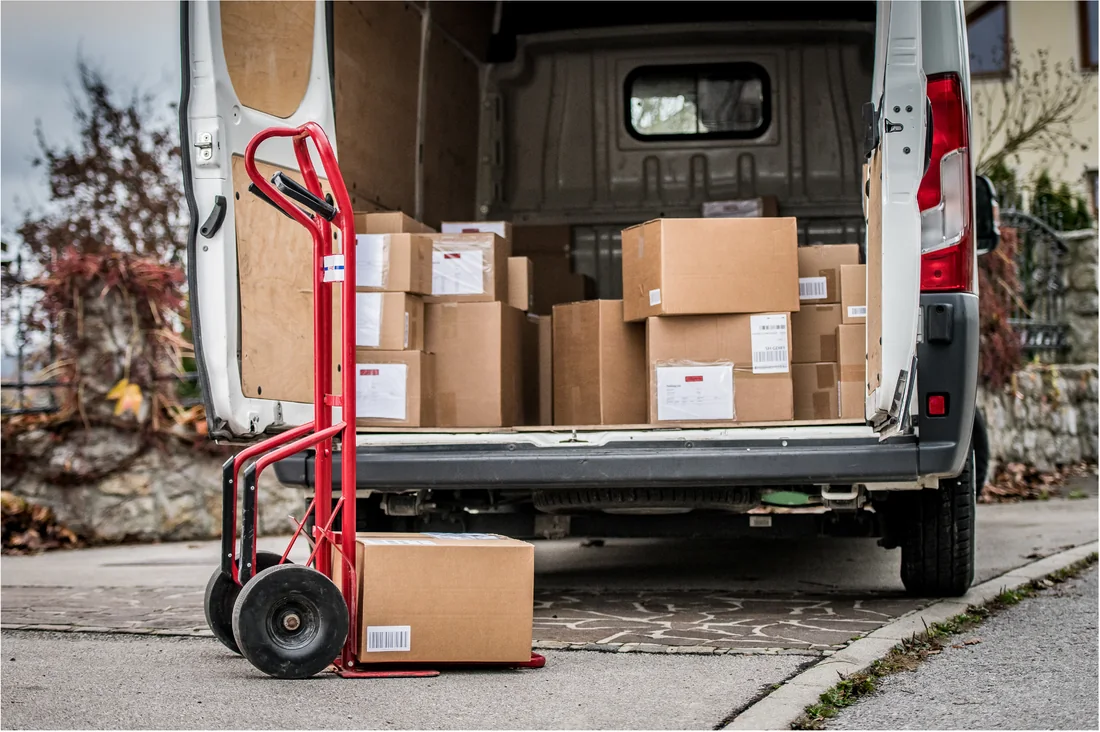 A loaded van with its rear doors open, a sack truck with a box standing in front of it